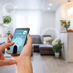 Mano interactuando con app "SMART HOME" en un smartphone, controlando una casa tecnológica moderna con luces y seguridad.